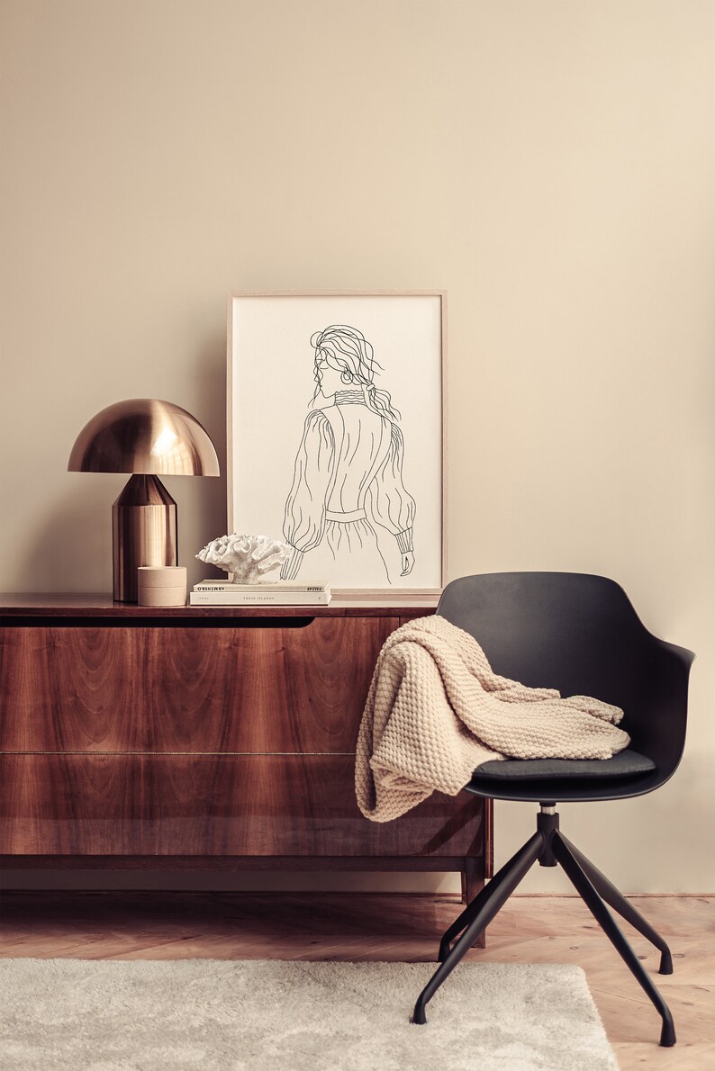 K&ouml;nnte beinhalten: Ein schwarzer Stuhl mit einer beigefarbenen Strickdecke dar&uuml;ber steht vor einer Kommode aus Holz. Eine goldene Lampe mit einem wei&szlig;en Schirm steht auf der Kommode neben einer gerahmten Schwarzwei&szlig;-Zeichnung einer Frau.