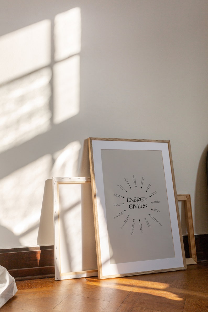 Puede incluir: Una impresi&oacute;n enmarcada con un fondo beige y texto negro que dice "ENERGY GIVERS" en un c&iacute;rculo con flechas que apuntan a palabras como "meditaci&oacute;n", "lectura", "baile" y "naturaleza". La impresi&oacute;n est&aacute; en un marco de madera claro y se apoya contra una pared blanca.