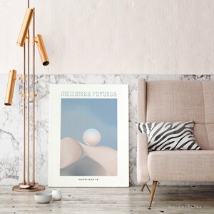 May include: A poster with the text "M&Eacute;MOIRES FUTURS" in blue lettering. The poster features a light blue sky with a white orb in the center. The orb is resting on a beige, rounded hill. The poster is framed and hanging on a white wall.