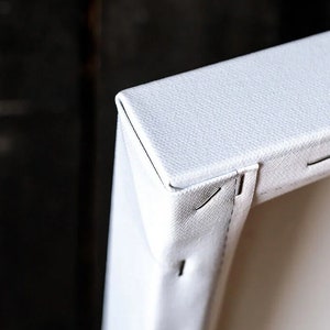May include: Close-up of the corner of a white canvas stretched over a wooden frame. The canvas is secured with staples.