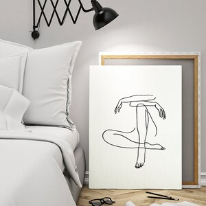 May include: A black and white line drawing of a person sitting with their legs crossed. The drawing is on a white canvas with a wooden frame. The canvas is leaning against a white wall.
