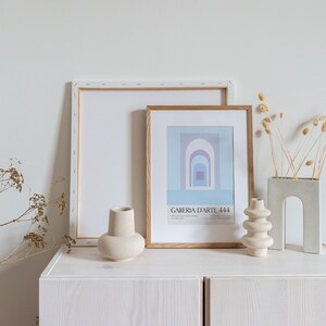 Peut inclure: Deux cadres, l'un avec un fond blanc et l'autre avec un fond bleu et des formes g&eacute;om&eacute;triques abstraites, sont expos&eacute;s sur un meuble blanc avec une texture en bois. Il y a trois vases en c&eacute;ramique, deux blancs et un gris, et des fleurs s&eacute;ch&eacute;es dans un vase gris.