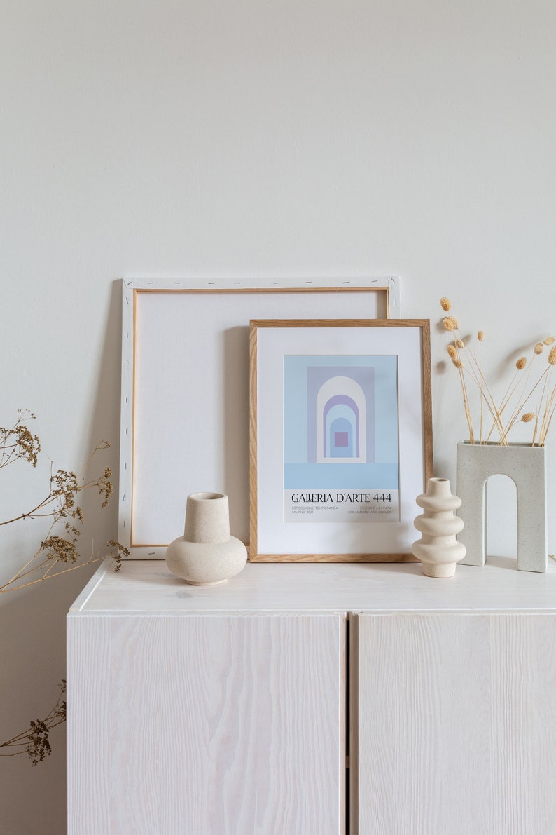 Peut inclure: Deux cadres, l'un avec un fond blanc et l'autre avec un fond bleu et des formes g&eacute;om&eacute;triques abstraites, sont expos&eacute;s sur un meuble blanc avec une texture en bois. Il y a trois vases en c&eacute;ramique, deux blancs et un gris, et des fleurs s&eacute;ch&eacute;es dans un vase gris.
