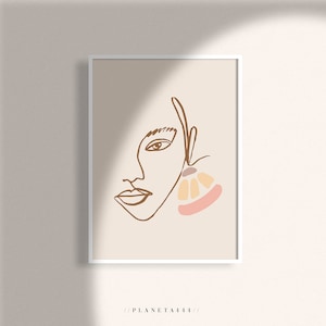 以下が含まれることがあります： ピンク、オレンジ、ブラウンの色合いの大きな抽象的なイヤリングをつけた顔のミニマルな線画プリント。背景は薄いベージュ色。プリントは白いフレームで額装されています。