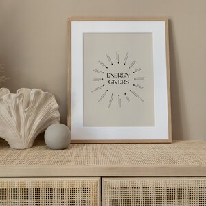 Puede incluir: Una impresi&oacute;n enmarcada con un fondo beige y texto negro que dice "ENERGY GIVERS" en un c&iacute;rculo con flechas que apuntan a palabras como "lectura", "meditaci&oacute;n", "sol" y "risa".