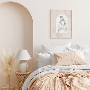 K&ouml;nnte beinhalten: Ein wei&szlig;es Bett mit einer beige gestrickten Wolldecke und wei&szlig;en Kissen. An der Wand &uuml;ber dem Bett h&auml;ngt ein gerahmtes Strichbild einer Frau in einem Kleid. Eine wei&szlig;e Lampe mit wei&szlig;em Schirm steht auf einem Korb-Beistelltisch neben dem Bett.
