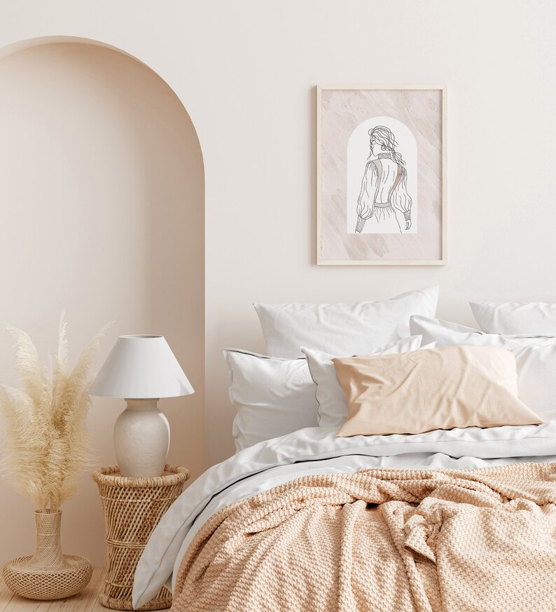 K&ouml;nnte beinhalten: Ein wei&szlig;es Bett mit einer beige gestrickten Wolldecke und wei&szlig;en Kissen. An der Wand &uuml;ber dem Bett h&auml;ngt ein gerahmtes Strichbild einer Frau in einem Kleid. Eine wei&szlig;e Lampe mit wei&szlig;em Schirm steht auf einem Korb-Beistelltisch neben dem Bett.