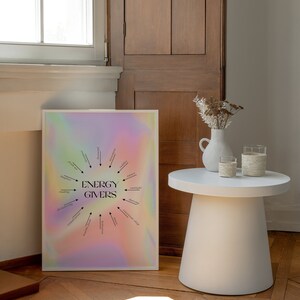 Puede incluir: Un p&oacute;ster enmarcado blanco con un fondo hologr&aacute;fico y texto negro que dice "ENERGY GIVERS" con un diagrama circular de actividades que dan energ&iacute;a, como leer, bailar y la luz del sol. El p&oacute;ster est&aacute; en un suelo de madera con una mesa auxiliar blanca y un jarr&oacute;n con flores secas.