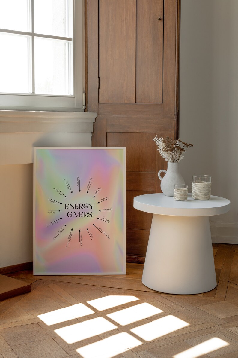 Puede incluir: Un p&oacute;ster enmarcado blanco con un fondo hologr&aacute;fico y texto negro que dice "ENERGY GIVERS" con un diagrama circular de actividades que dan energ&iacute;a, como leer, bailar y la luz del sol. El p&oacute;ster est&aacute; en un suelo de madera con una mesa auxiliar blanca y un jarr&oacute;n con flores secas.