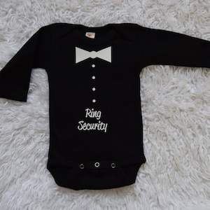 May include: Black baby bodysuit with long sleeves, featuring a white bow tie, faux buttons, and the text "Ring Security" in white. The bodysuit has snap closures at the bottom and is designed for infants.