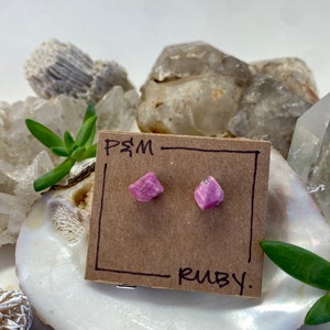 Puede incluir: Un par de pendientes de rubí rosa en una tarjeta marrón con el texto "PSM" y "RUBY" escrito en ella. Los pendientes se muestran en una concha blanca.