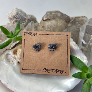 May include: A pair of silver stud earrings with a black geode design. The earrings are displayed on a brown card with the text "PSM - GEODE."