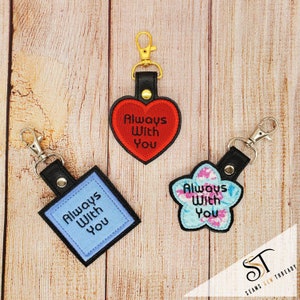 May include: Three keychains with the text "Always With You" embroidered on them. The keychains are in the shapes of a heart, a square, and a flower. The heart is red, the square is blue, and the flower is blue and pink.