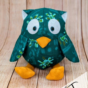 May include: A teal blue owl plush toy with a green and white patterned fabric. The owl has large white eyes with black pupils, an orange beak, and orange feet.