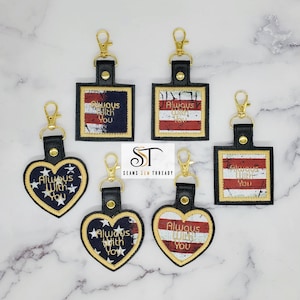 May include: Five keychains with a red, white, and blue American flag design. The keychains are in the shape of a square and a heart. The text "Always With You" is embroidered on each keychain.
