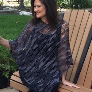May include: A gray crochet poncho with a striped pattern. The poncho is worn by a person sitting on a wooden bench.