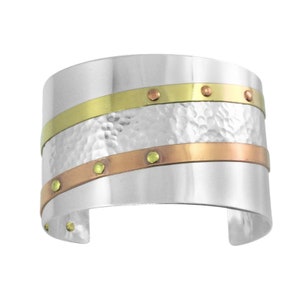 May include: Wide silver cuff bracelet with hammered texture and two bands of gold and copper-colored metal. The bands are accented with small, evenly spaced rivets. The bracelet has an open design.