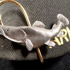 May include: A silver-colored fish figurine with detailed scales and fins, attached to a gold-colored hook. The fish is mounted on a black and green baseball cap.