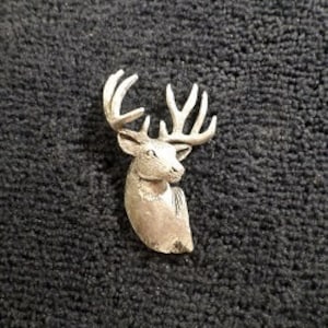 Könnte beinhalten: Eine silberfarbene Metall-Hirschkopfnadel mit einem detaillierten Design. Der Hirsch hat große Geweihe und eine realistische Optik.