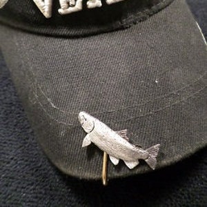 Puede incluir: Un pin de pescado de color plateado con un cierre de aguja, fijado a una gorra de b&eacute;isbol negra con la palabra "Veteran" bordada en la parte delantera.