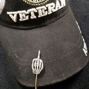 May include: Black cap with "VETERAN" embroidered in white, and a U.S. Army emblem. A silver-colored pin shaped like a skeletal hand giving the middle finger is attached to the cap.