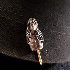 May include: A silver-toned hat pin featuring a detailed sculpted bust of a woman with long hair. The pin has a gold-toned stem.