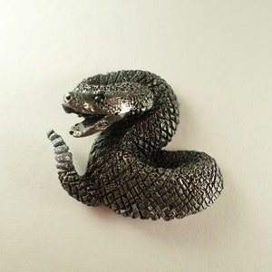 May include: A silver-colored pewter figurine of a coiled rattlesnake with its head raised and mouth open. The snake has a detailed, textured surface.