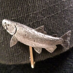 Puede incluir: Un broche de corbata de pez de esta&ntilde;o plateado. El pez est&aacute; detallado con escamas y aletas. El broche de corbata est&aacute; unido a un alfiler dorado.