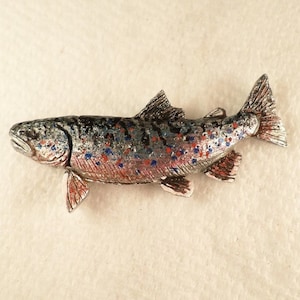 Puede incluir: Una figurita de pez trucha de color plateado con manchas pintadas de rojo y azul. El pez est&aacute; detallado con aletas y escamas.