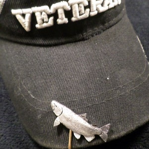 Puede incluir: Una gorra de b&eacute;isbol negra con la palabra "VETERAN" bordada en la parte delantera. Un alfiler de sombrero de pescado plateado est&aacute; sujeto a la gorra.