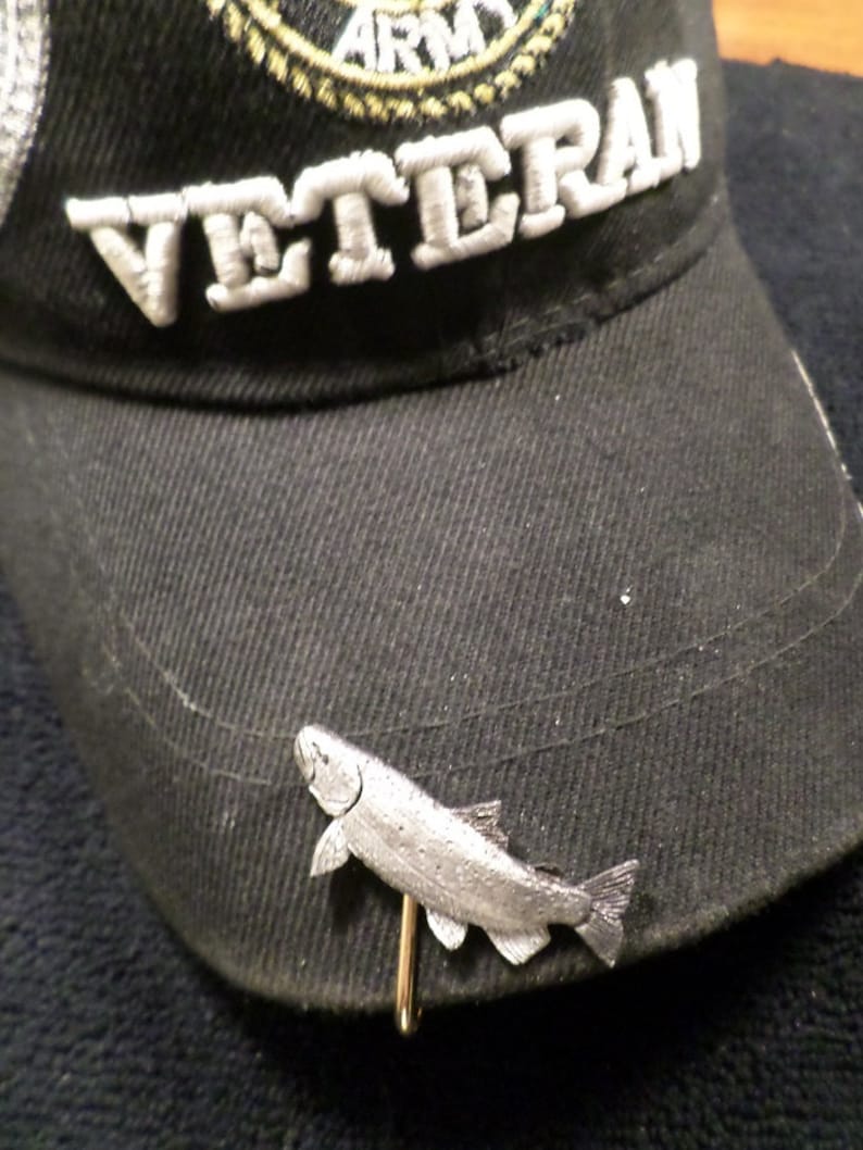 Puede incluir: Una gorra de b&eacute;isbol negra con la palabra "VETERAN" bordada en la parte delantera. Un alfiler de sombrero de pescado plateado est&aacute; sujeto a la gorra.