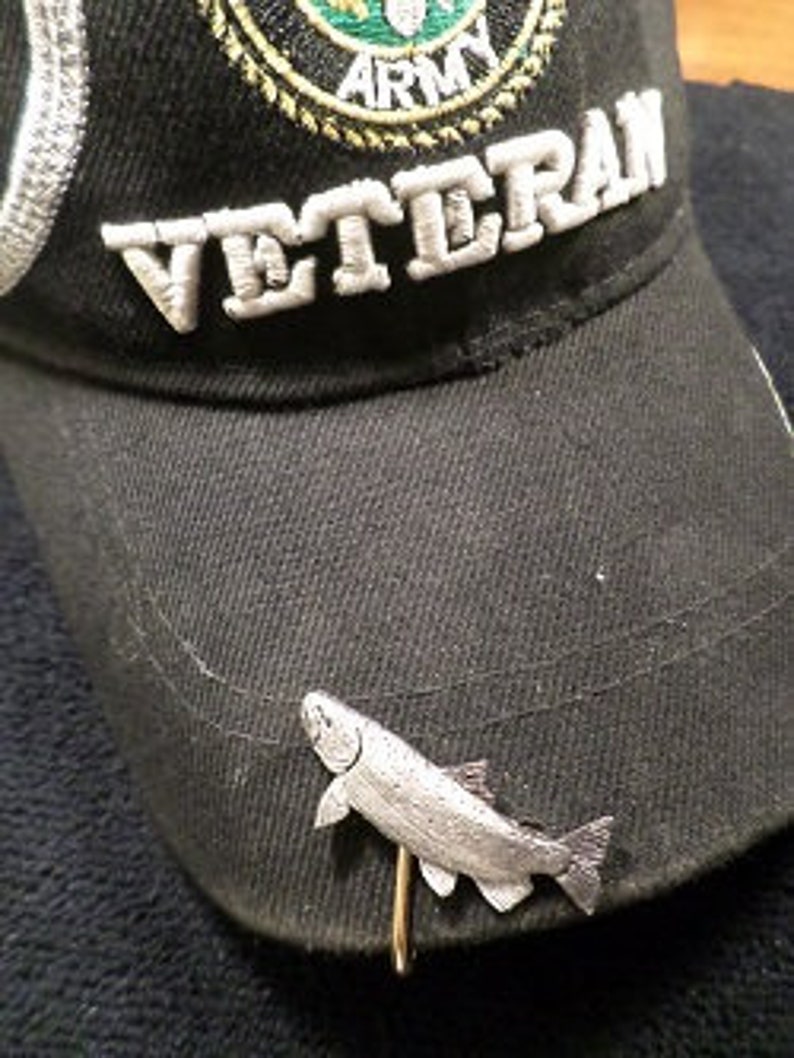 Puede incluir: Un pin de pescado de color plateado con un cierre de aguja, fijado a una gorra de b&eacute;isbol negra con la palabra "Veteran" bordada en la parte delantera.