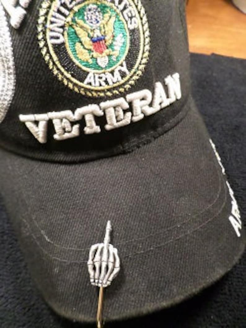 May include: Black cap with "VETERAN" embroidered in white, and a U.S. Army emblem. A silver-colored pin shaped like a skeletal hand giving the middle finger is attached to the cap.