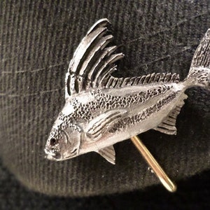 May include: A silver-colored fish pin with a detailed design. The fish has a long, thin body with a large dorsal fin and a small tail fin. The pin is attached to a gold-colored pin back.