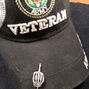 May include: Black baseball cap with "VETERAN" embroidered in white. Features a U.S. Army emblem and a silver-colored pin of a skeleton hand giving the middle finger.