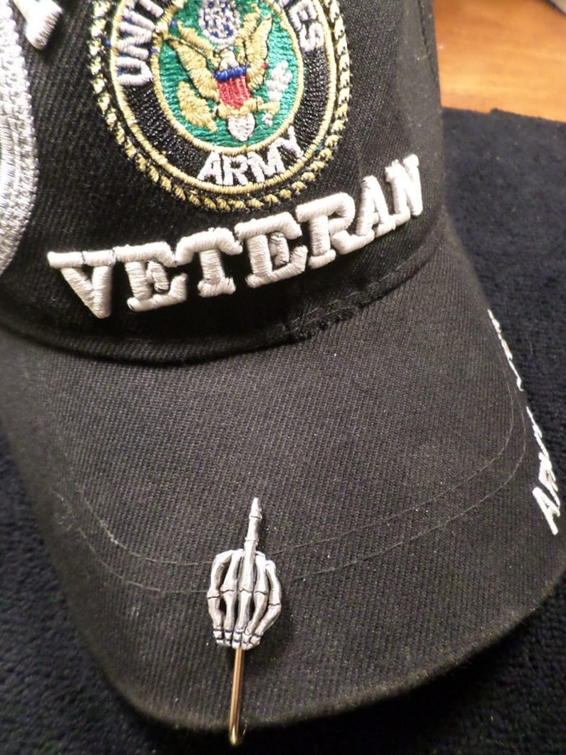 May include: Black baseball cap with "VETERAN" embroidered in white. Features a U.S. Army emblem and a silver-colored pin of a skeleton hand giving the middle finger.