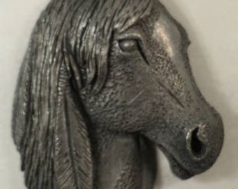 May include: A detailed, metallic sculpture of a horse's head. The sculpture is a muted silver color, with visible texture and detail in the mane, eye, and muzzle. The horse's head is angled slightly to the right.