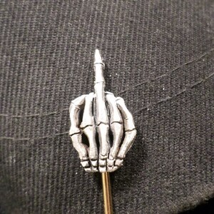 May include: A silver-colored lapel pin shaped like a skeleton hand giving the middle finger. The pin has a gold-colored pin backing and is set against a dark gray textured fabric.