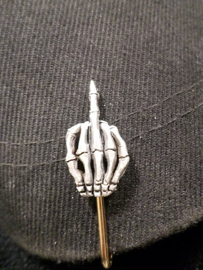 May include: A silver-colored lapel pin shaped like a skeleton hand giving the middle finger. The pin has a gold-colored pin backing and is set against a dark gray textured fabric.