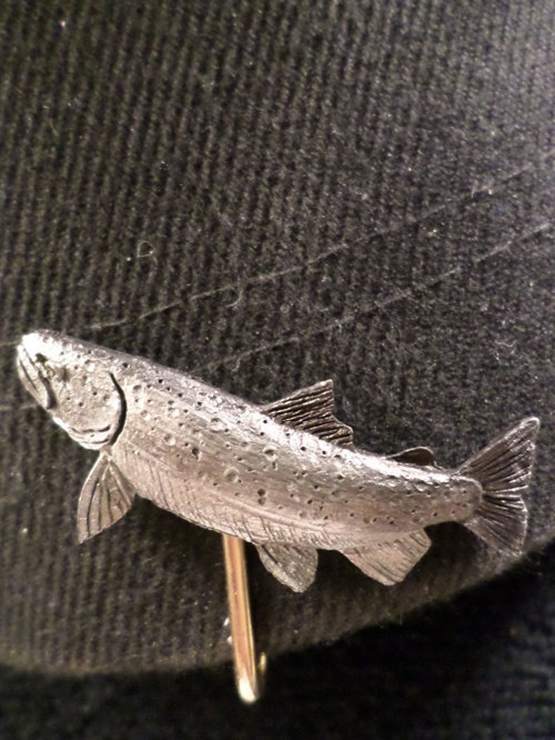 Puede incluir: Un broche de corbata de pez de esta&ntilde;o plateado. El pez est&aacute; detallado con escamas y aletas. El broche de corbata est&aacute; unido a un alfiler dorado.