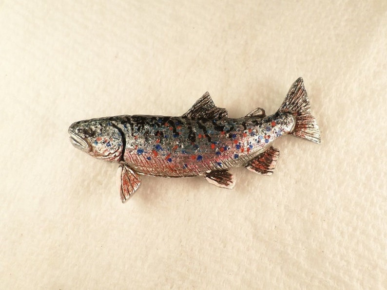 Puede incluir: Una figurita de pez trucha de color plateado con manchas pintadas de rojo y azul. El pez est&aacute; detallado con aletas y escamas.