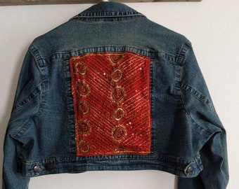 May include: A blue denim jacket with a cropped hem and long sleeves. The back of the jacket features a red and gold sequined patch.
