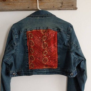 May include: A blue denim jacket with a cropped hem and long sleeves. The back of the jacket features a red and gold sequined patch.
