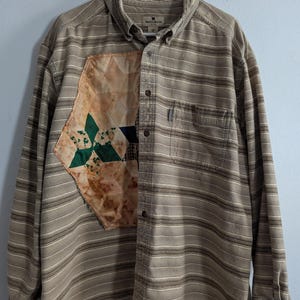 May include: A brown and white striped long-sleeved shirt with a patchwork design on the left side. The shirt has a button-up front and a pocket on the left chest. The label on the shirt reads "Woolrich".