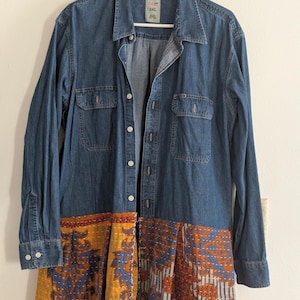 May include: A long denim shirt dress with a button-down front, two chest pockets, and long sleeves. The bottom half features a patchwork design in shades of gold, orange, and blue. The dress is hanging on a white hanger.