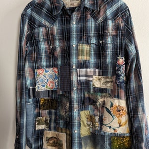 May include: A blue and brown plaid flannel shirt with various fabric patches sewn on. The patches feature floral patterns, animal prints, and landscapes. The shirt has a button-down front and long sleeves. The label reads "Urban Pipeline" and "Large".