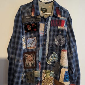 May include: Blue plaid shirt with various fabric patches sewn on. The patches are in different colors and patterns, including floral, geometric, and solid colors. The shirt has a button-down front and long sleeves with red cuffs.