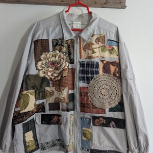 May include: A gray jacket with a zipper closure and a drawstring at the bottom. The jacket is decorated with various fabric patches in different colors and patterns, including floral, plaid, and lace.