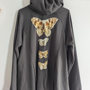 May include: A dark gray hooded sweatshirt with a long, flowing design. The back of the sweatshirt features four embroidered butterflies in shades of gold and brown.