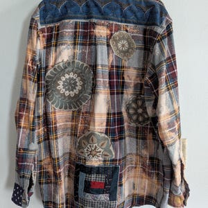May include: A plaid flannel shirt in shades of brown, burgundy, and gray, with decorative patches. The shirt features a blue patterned yoke and several circular and rectangular patches with intricate designs, adding a unique, handcrafted touch to the garment.